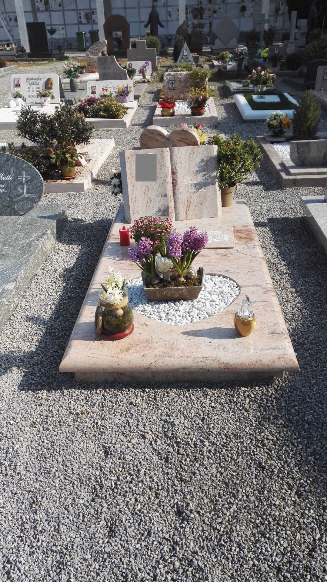 Tomba in granito rosa chiaro con stele a libro aperto e aiuola a cuore in ciottoli bianchi