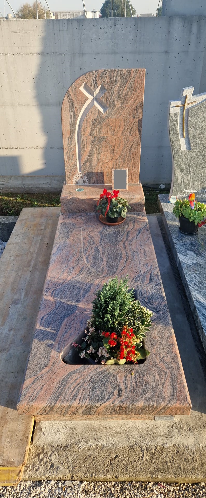 Tomba in granito rosa venato con stele ad arco, croce in rilievo e aiuola fiorita